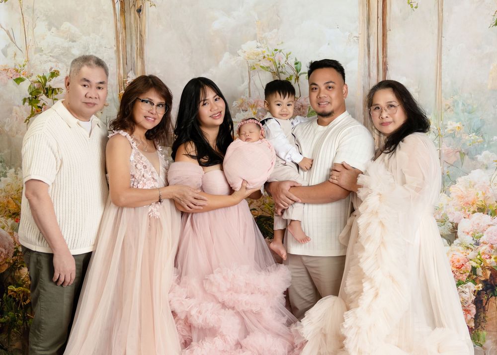 Multi-generational Asian family posing in elegant gowns with newborn baby in floral studio setting.