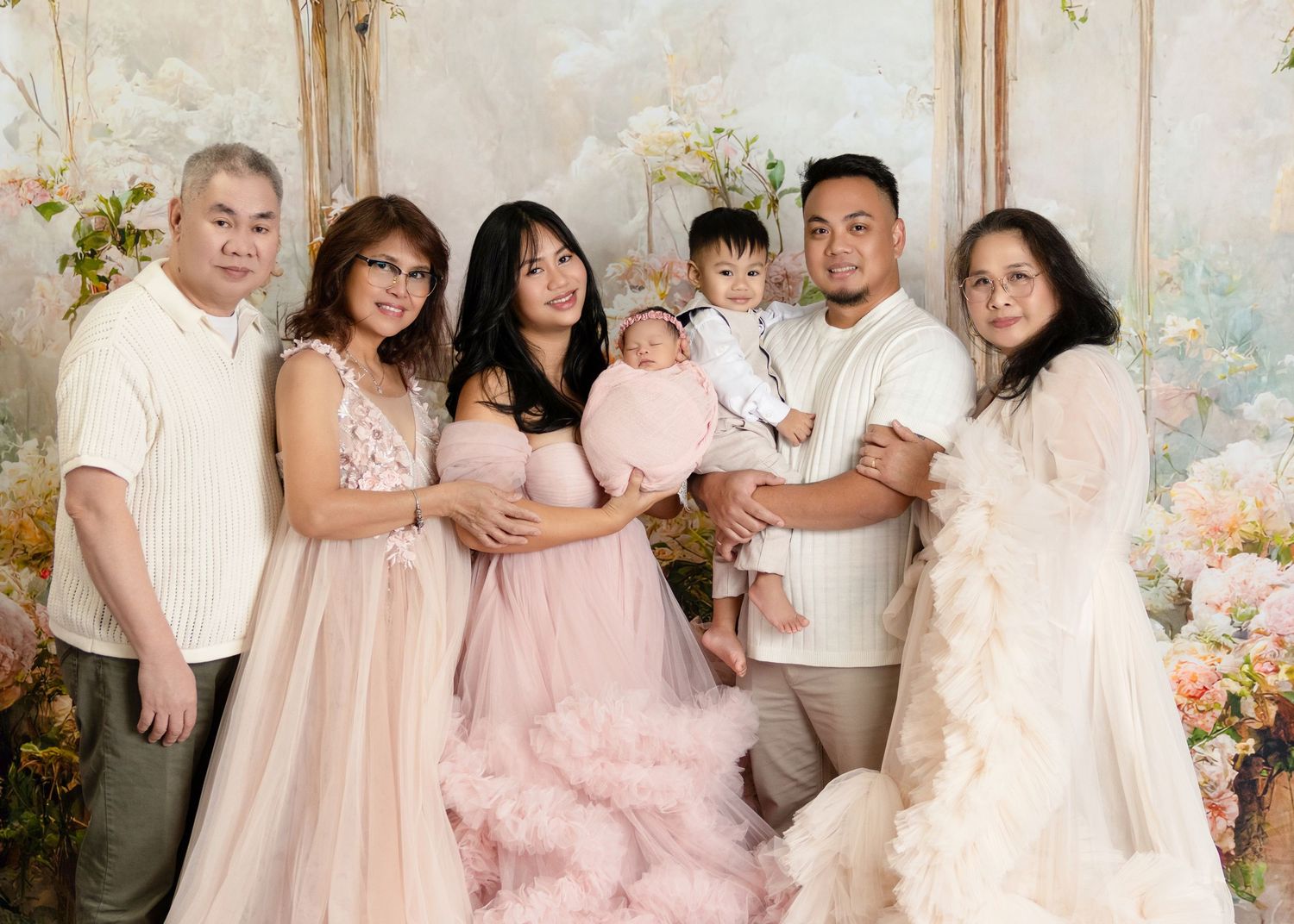 Multi-generational Asian family posing in elegant gowns with newborn baby in floral studio setting.