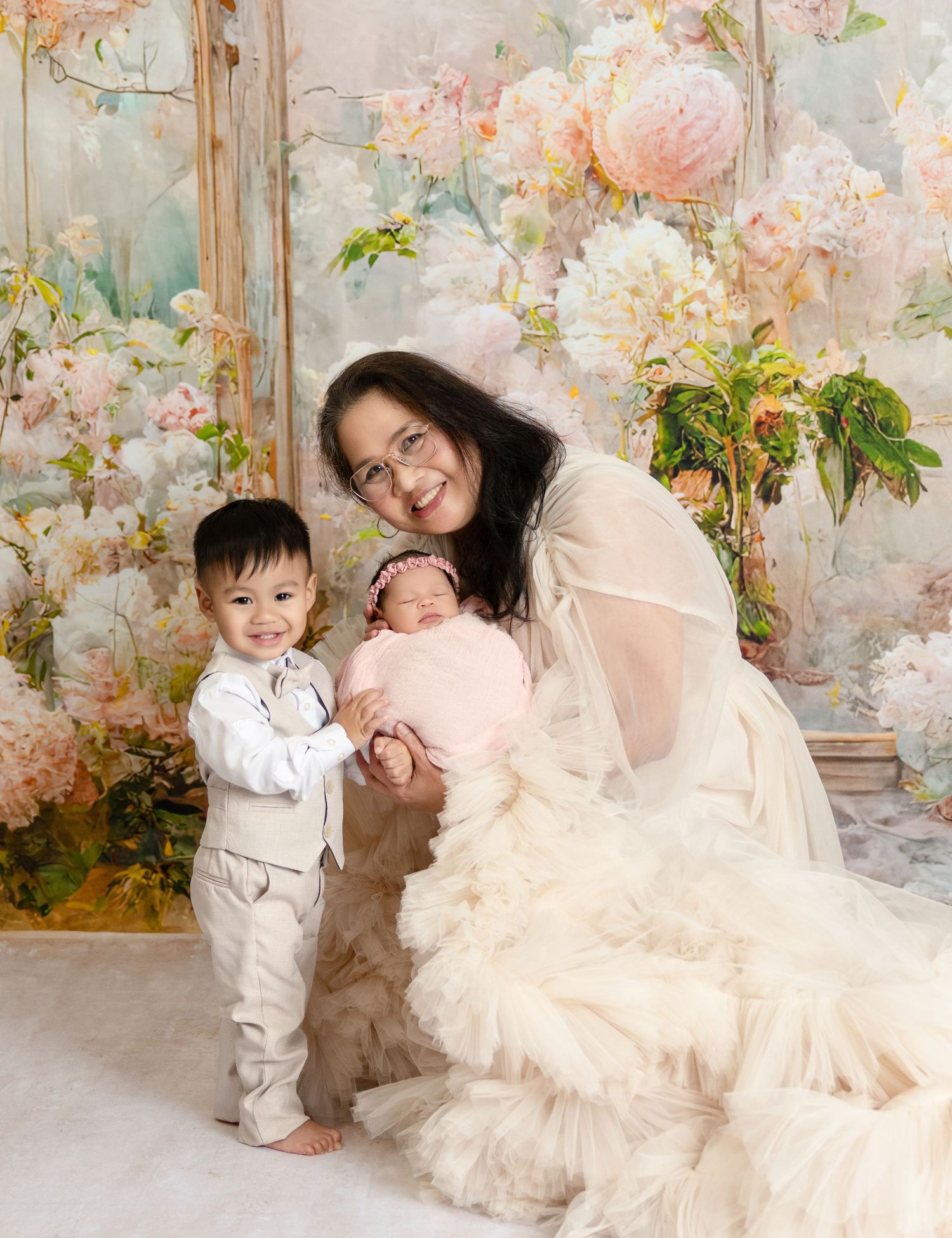 Mother in sheer gown sits with toddler son and swaddled newborn baby in floral studio portrait.