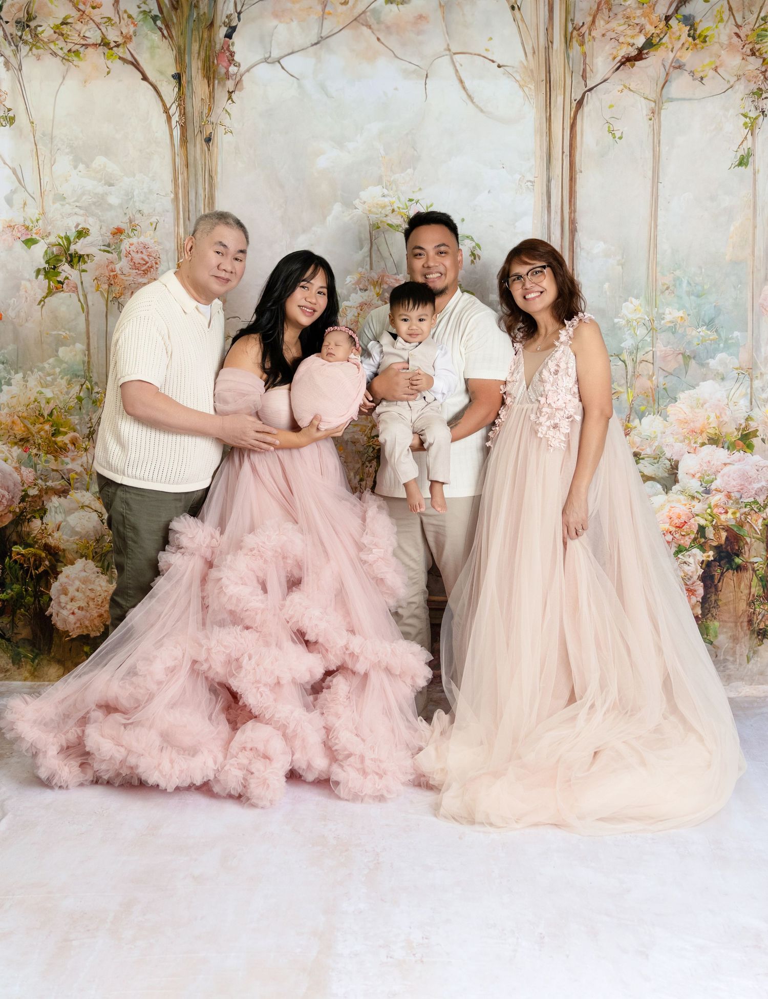 Asian family of five in elegant attire pose with newborn and toddler in floral studio setting.