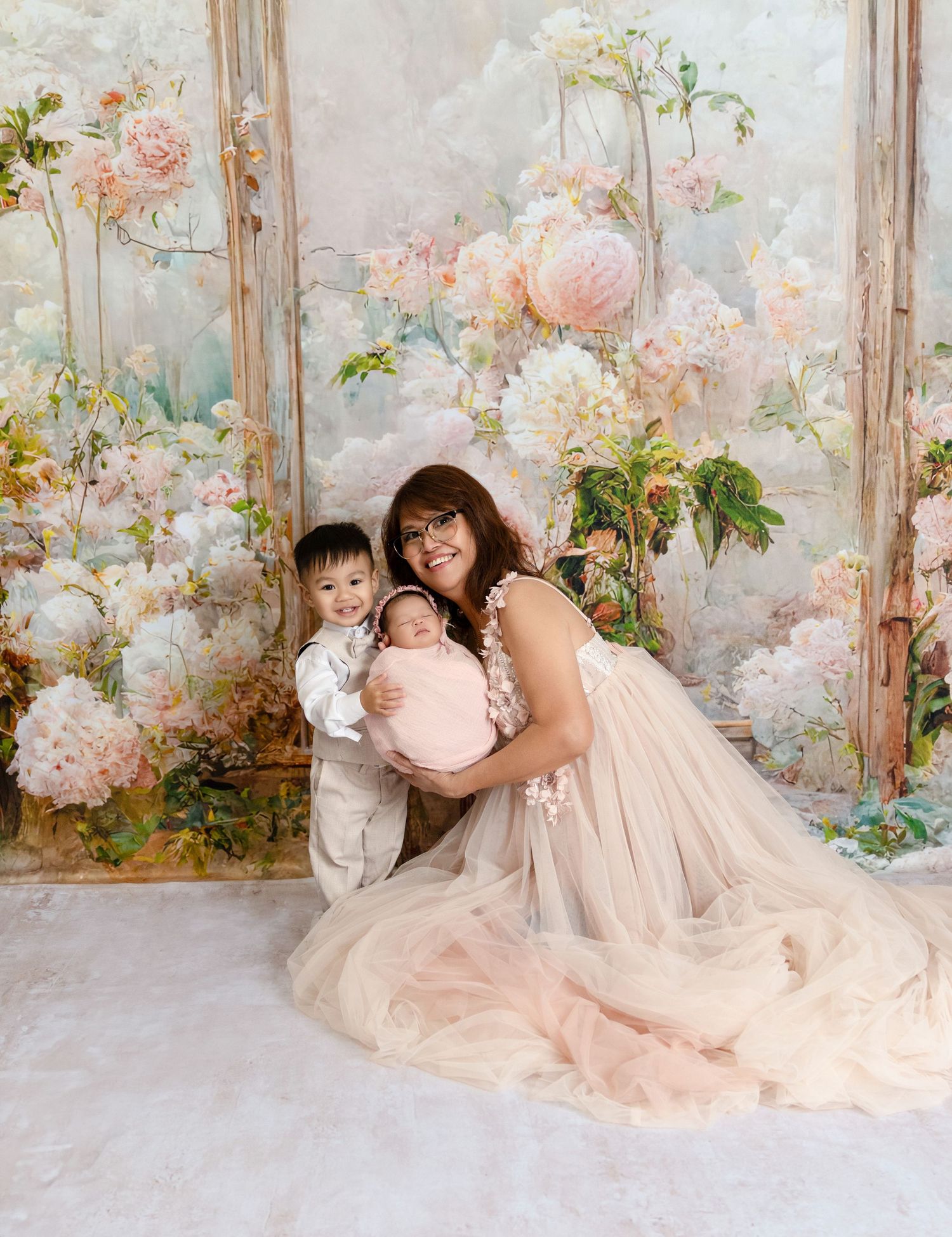 Mother in blush tulle gown sits with toddler son and swaddled newborn in floral studio portrait.