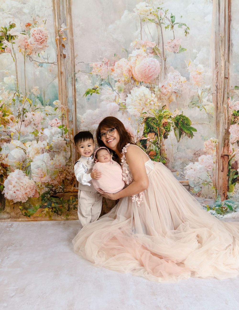Mother in blush tulle gown sits with toddler son and swaddled newborn in floral studio portrait.