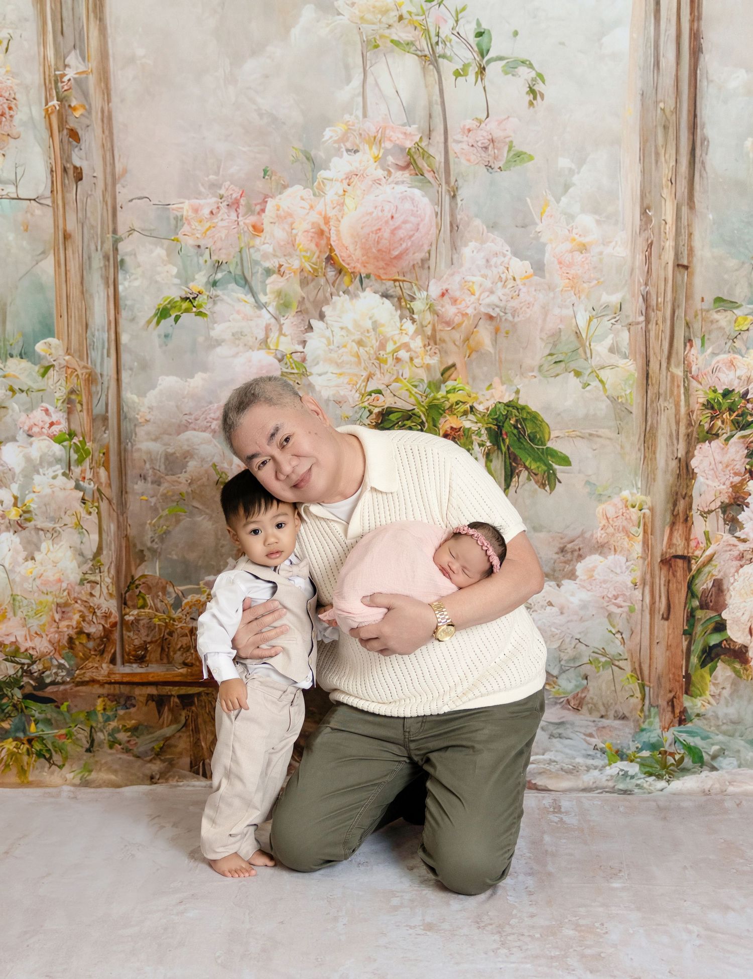 Grandfather kneels beside toddler grandson while holding swaddled newborn in a floral studio portrait.