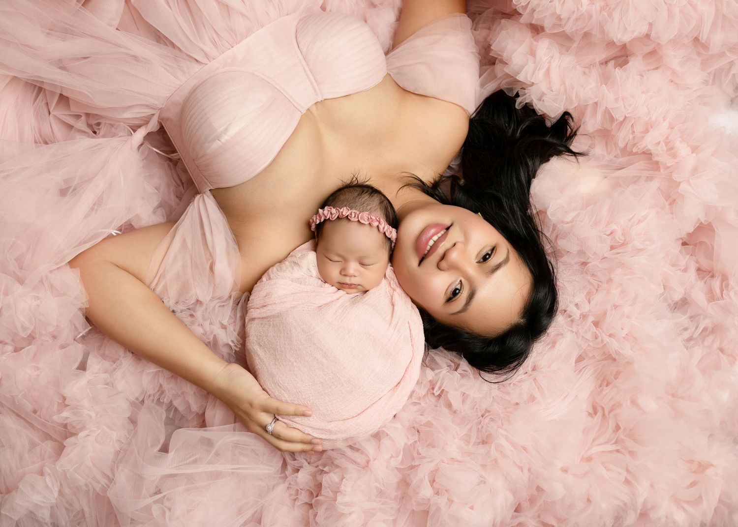 Mother in pink lingerie lying on pink tulle with sleeping newborn baby girl wrapped in pink.