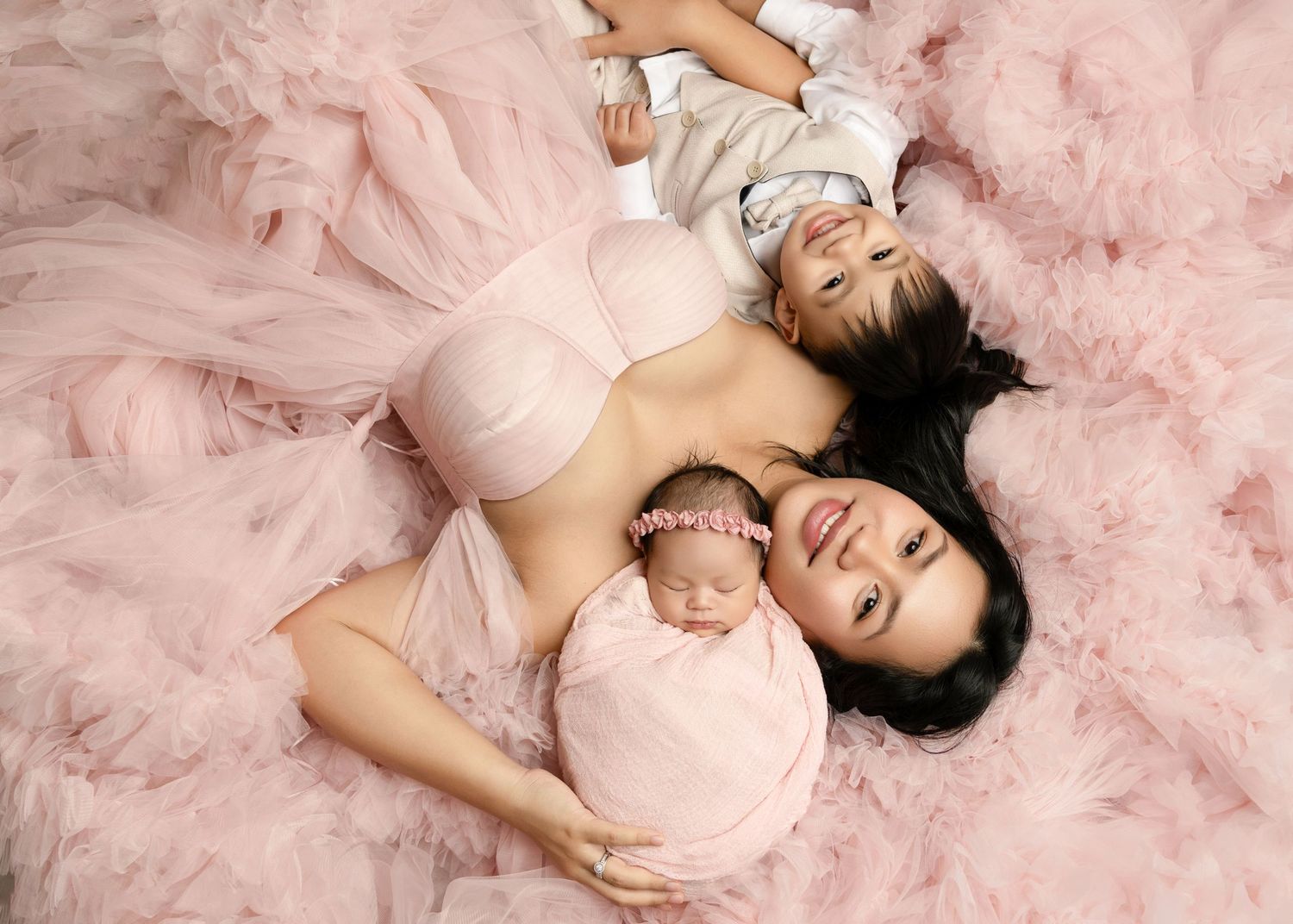 Mother in pink lingerie lies on pink tulle with toddler and newborn baby girl.