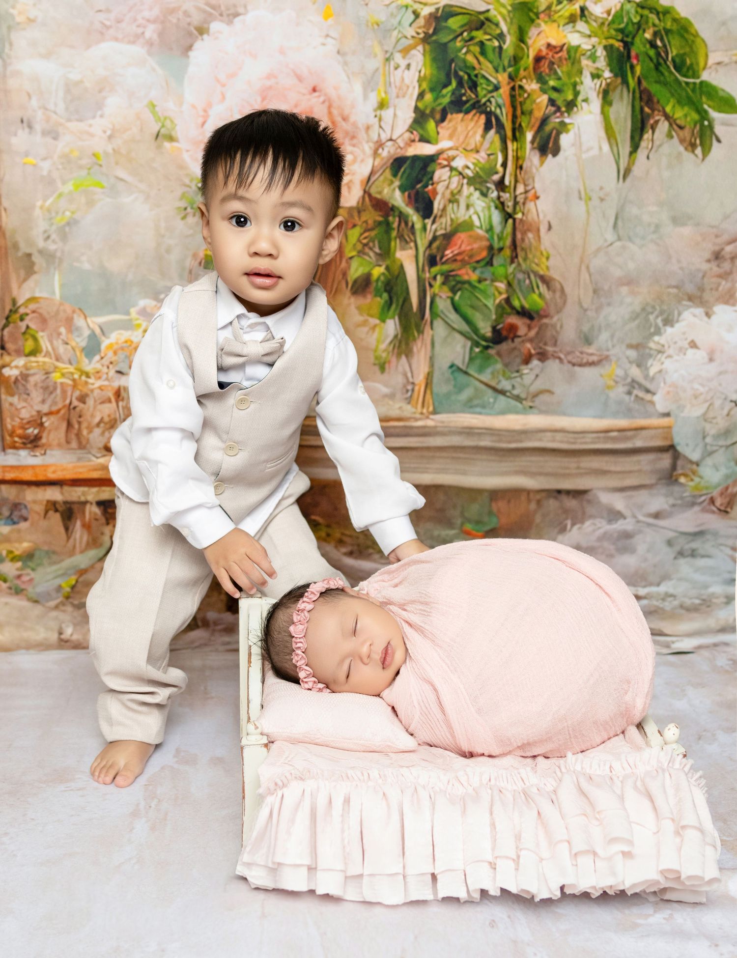 Toddler boy in tan suit poses with sleeping newborn sister on small pink ruffled bed.