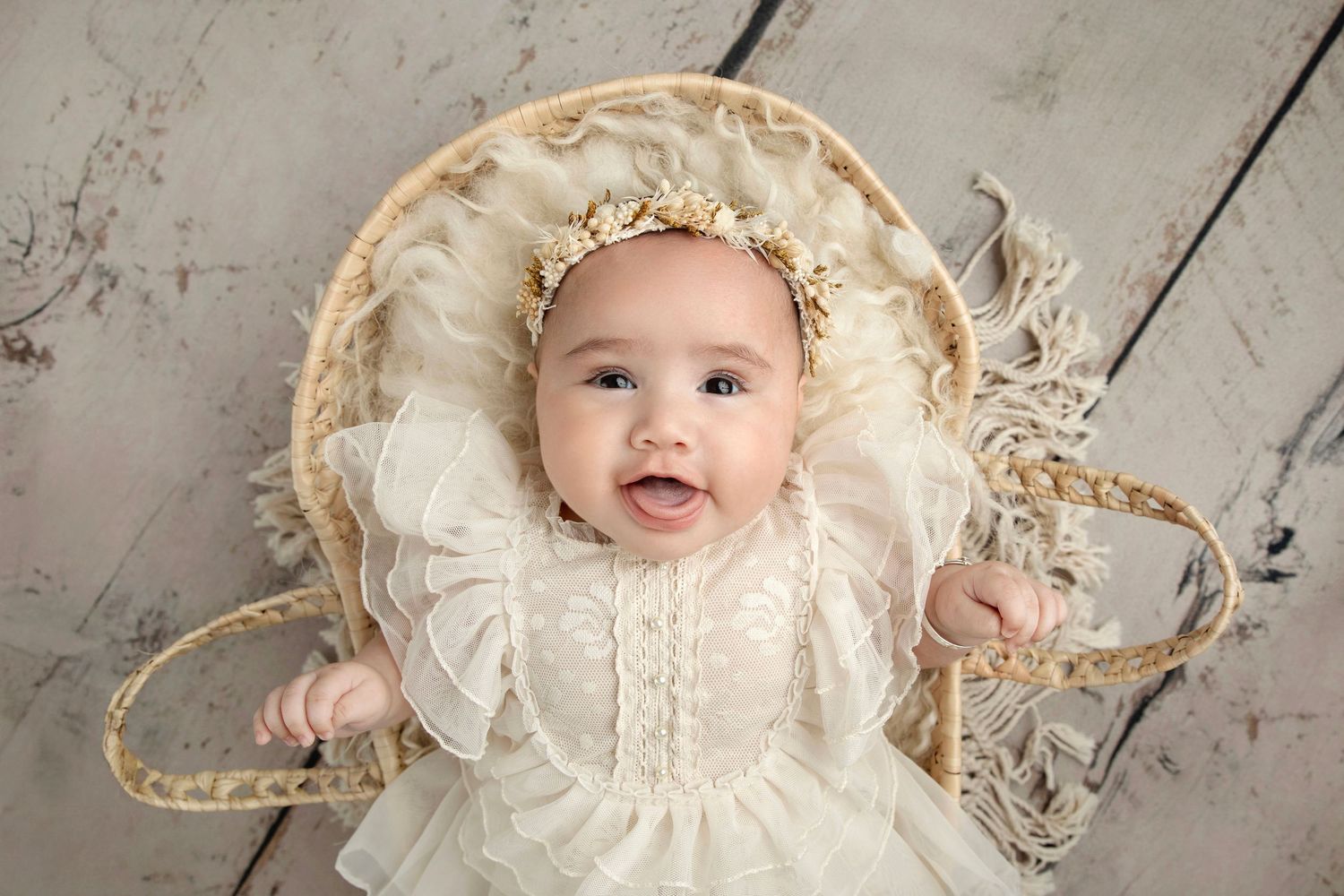 Smiling baby in white lace dress and gold crown headband lying in a wicker basket on a rustic wood floor.