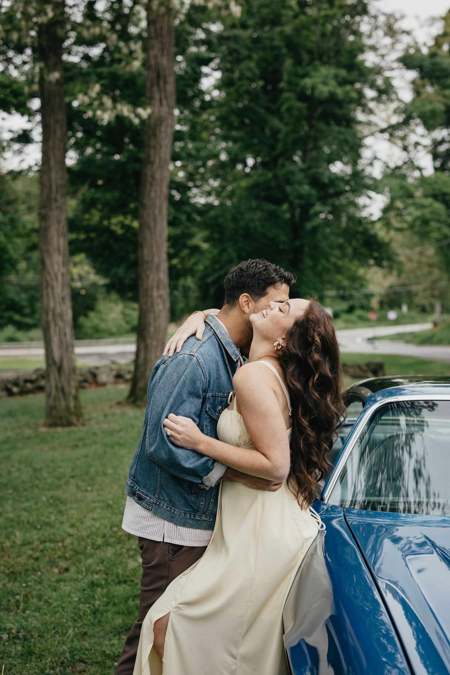 Couple sharing a romantic kiss beside a classic blue car surrounded by lush green trees outdoors.
