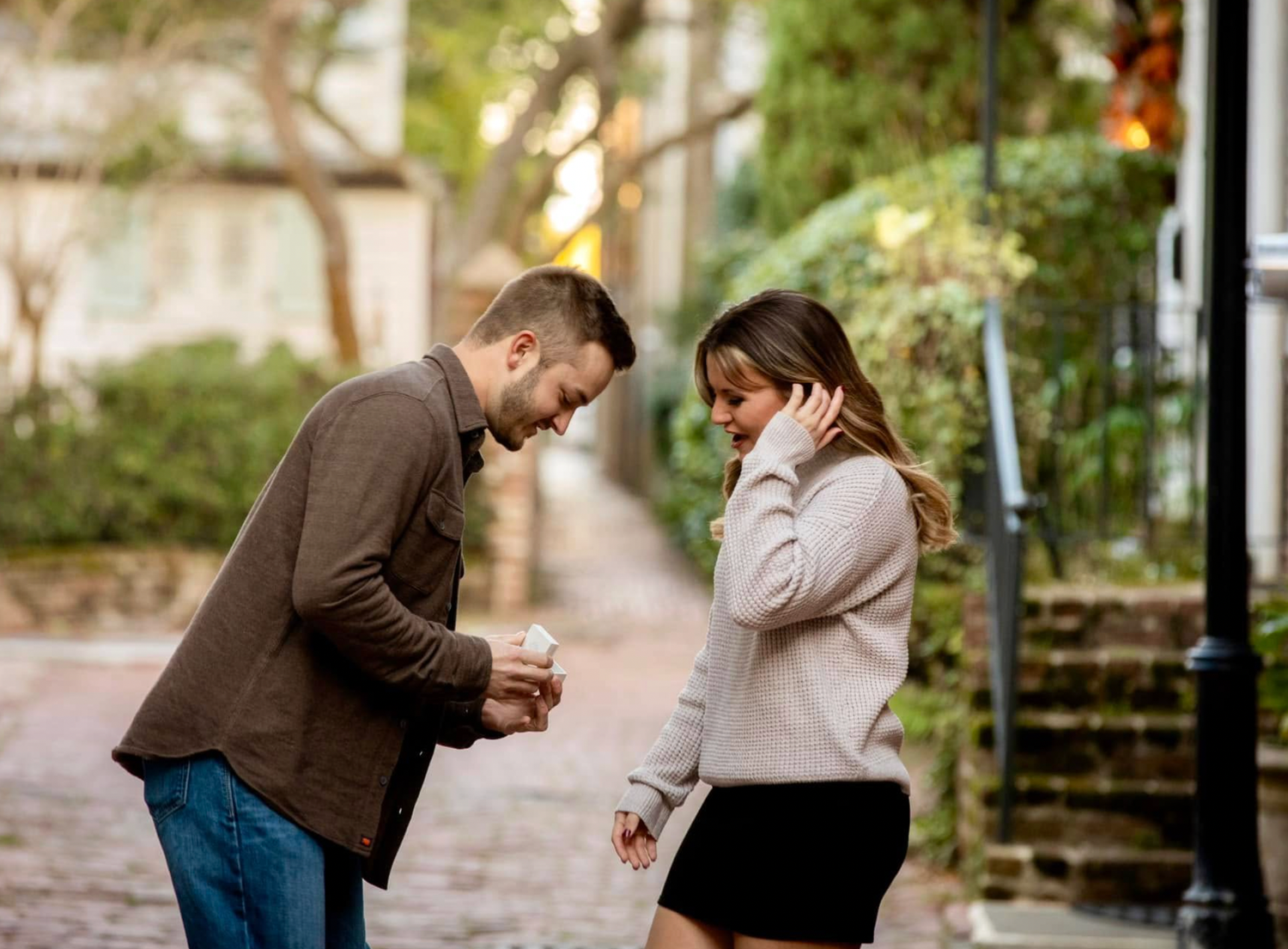 Couple sharing a romantic moment on a charming cobblestone street, man presenting a gift to smiling woman outdoors.
