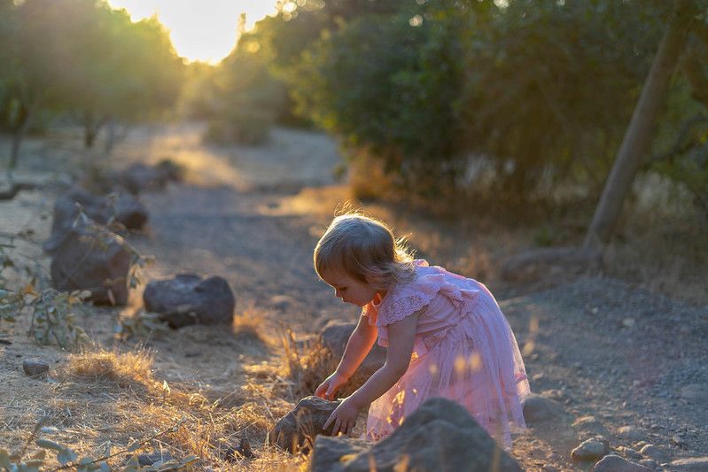 Child in pink dress plays with dirt on a dusty trail during golden hour sunset.