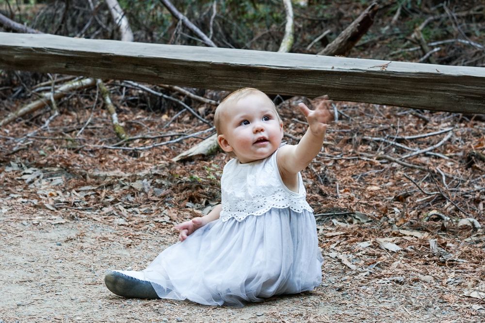 Baby in light blue dress sits on ground in outdoor setting while pointing up at something off camera.