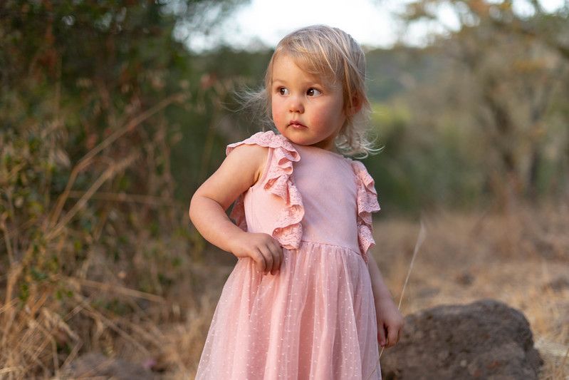 A small child wearing a light pink ruffle dress stands outdoors in golden sunlight against a blurred natural background.