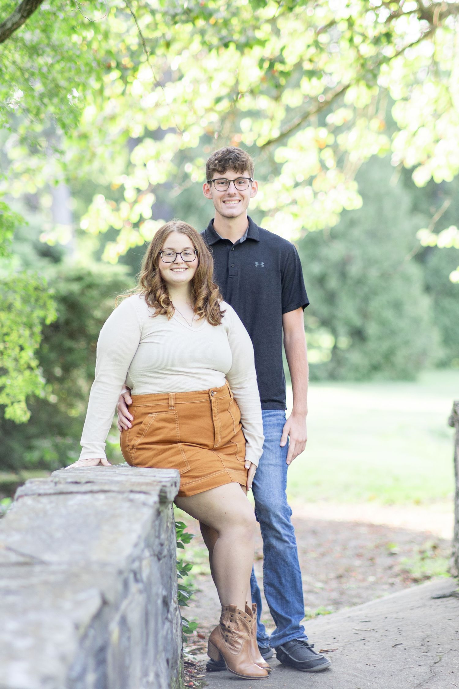 A young couple smiling and posing outdoors near a stone wall surrounded by lush green trees.