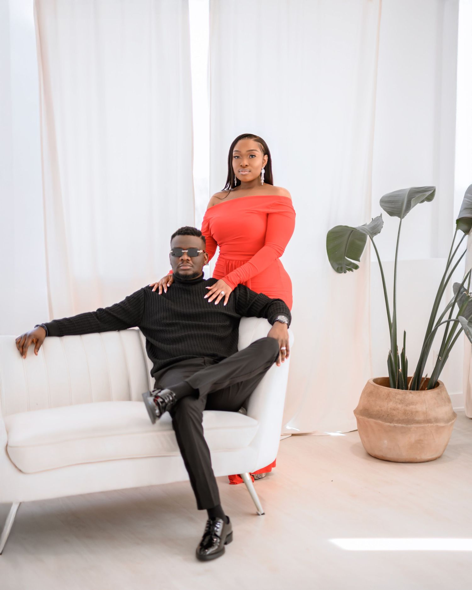 A stylish couple poses in a bright studio, man in black seated on white sofa, woman in red off-shoulder dress standing behind.
