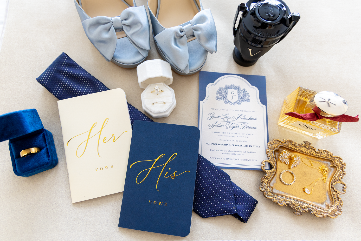 Flat lay of wedding details including blue vow books, invitation suite, rings, blue heels, and gold accessories.
