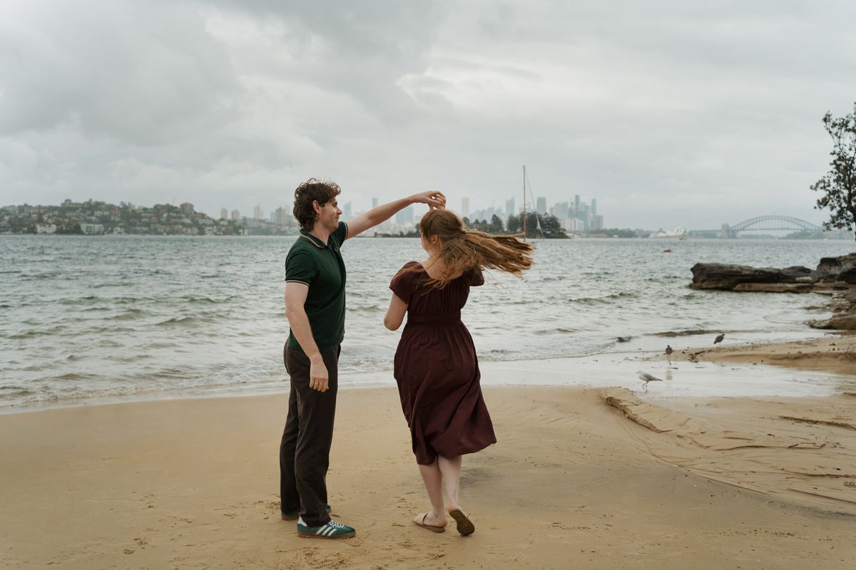 Couple dancing on sandy Milk beach with cloudy sky and  Sydney city skyline across the water in background during engagement photoshoot