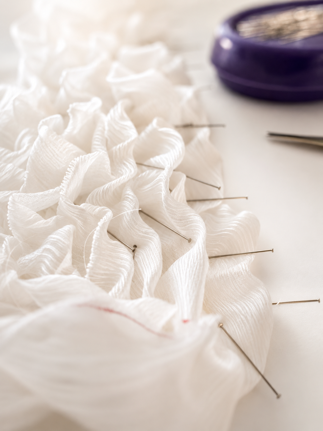 White ruffled fabric pinned with straight pins on a sewing surface, with a purple pincushion in the background.