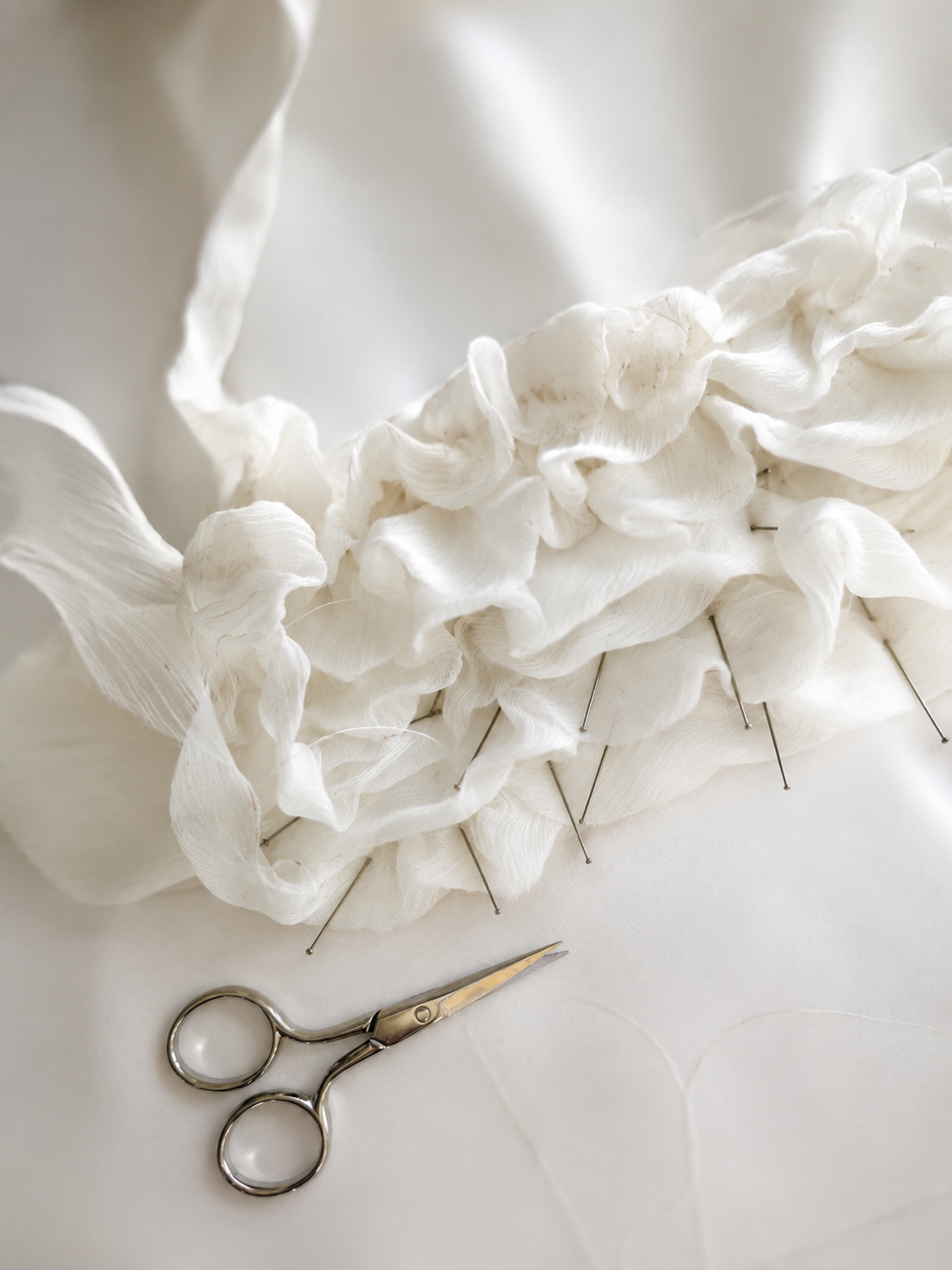 White ruffled fabric pinned to cloth with small gold scissors nearby on a white surface.