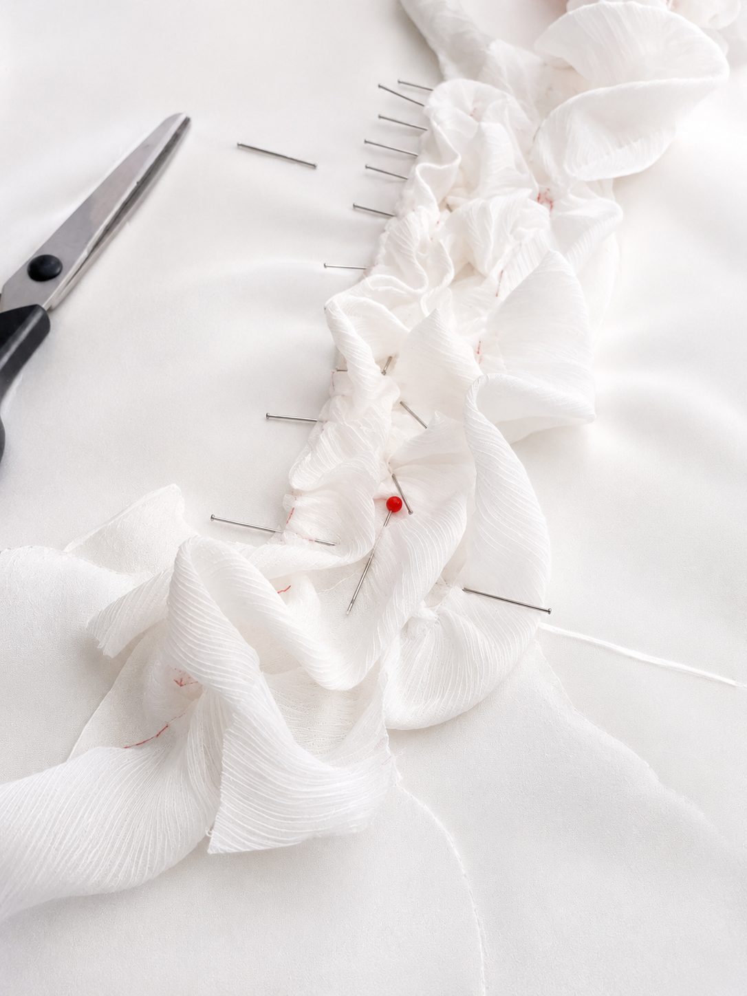 White fabric being pinned and gathered on a surface with sewing pins and scissors nearby.