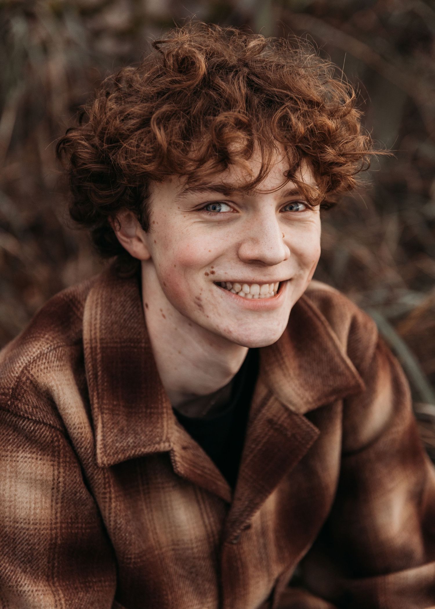 Smiling young man with curly red hair wearing a brown plaid jacket outdoors.