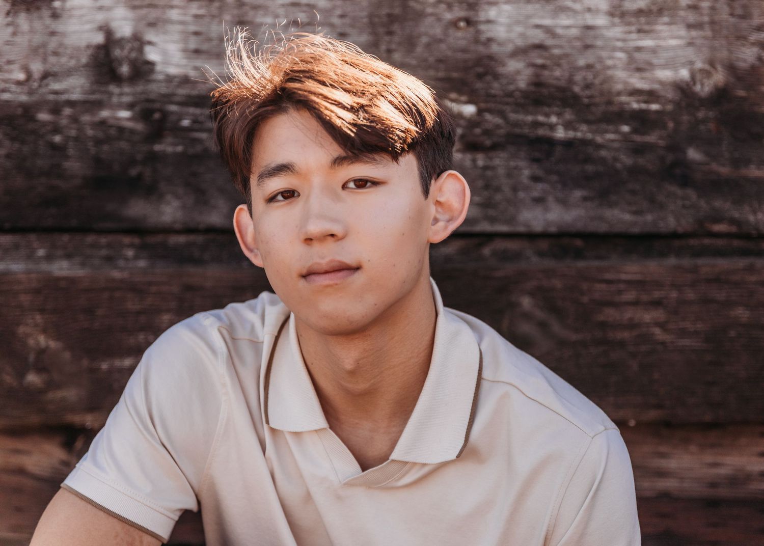 Young Asian man in a beige polo shirt poses outdoors against a rustic wooden wall in warm sunlight.