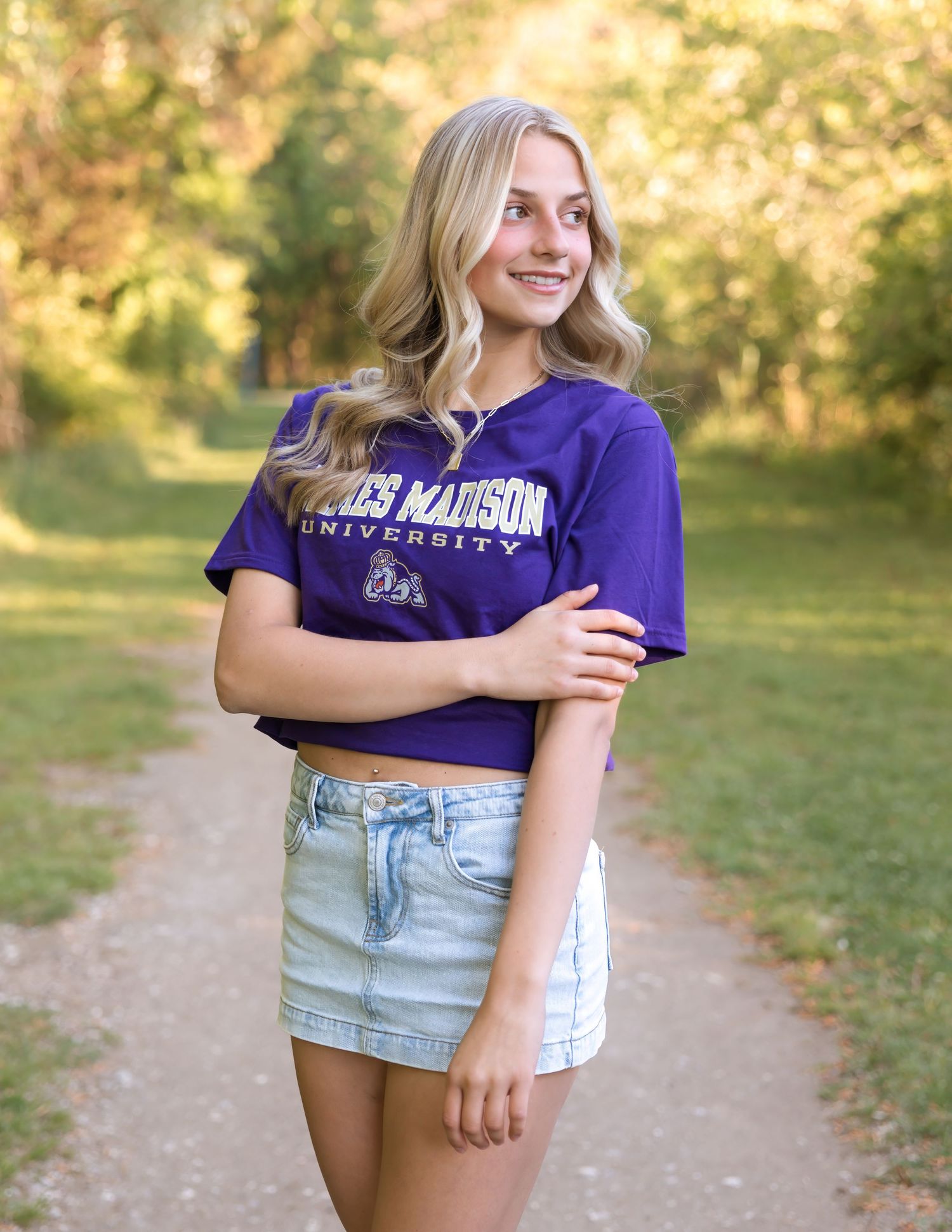 Smiling blonde woman wearing a purple James Madison University crop top and denim skirt standing on a tree-lined path.