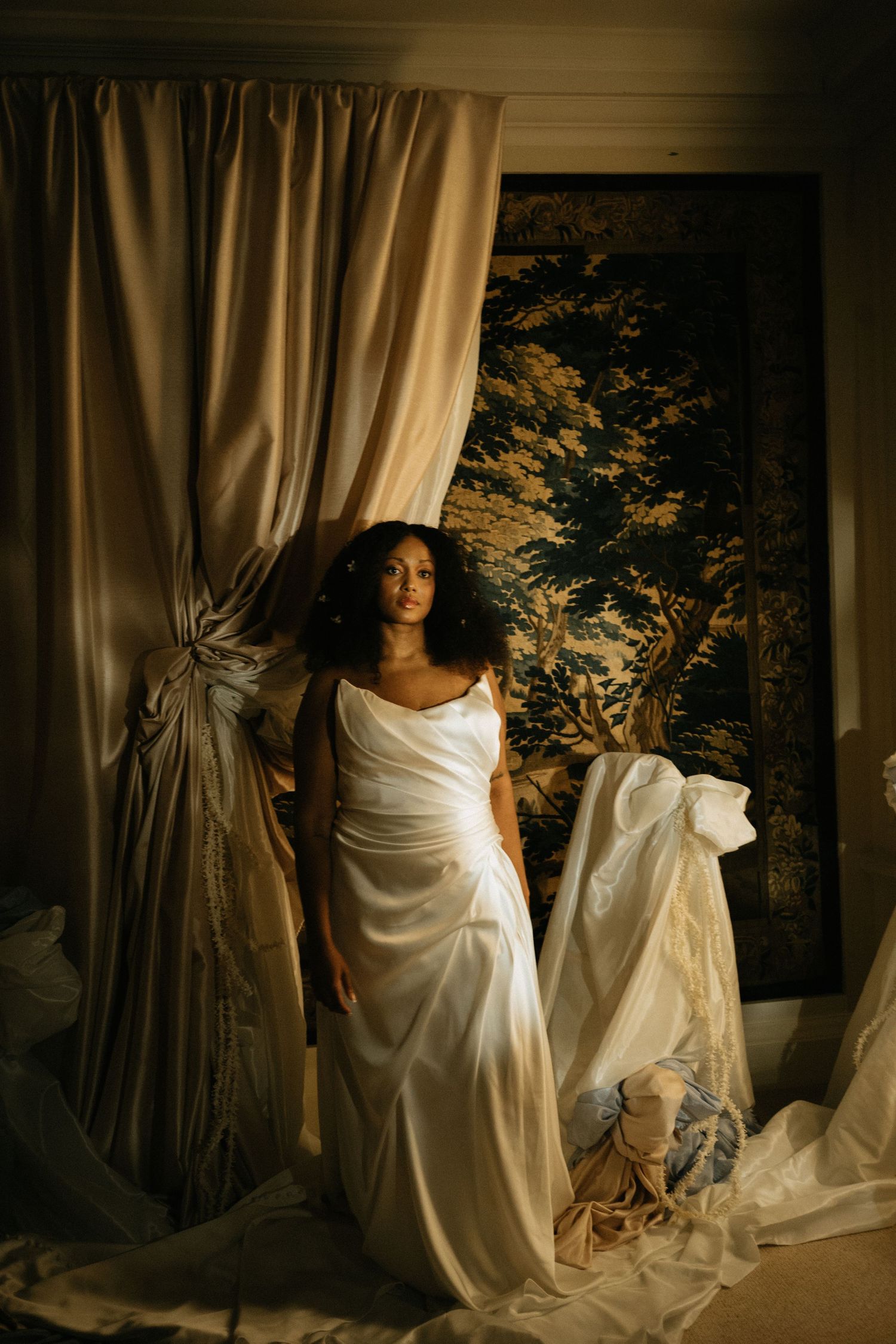 Woman in white satin gown standing between draped curtains before a scenic tapestry in moody golden light.