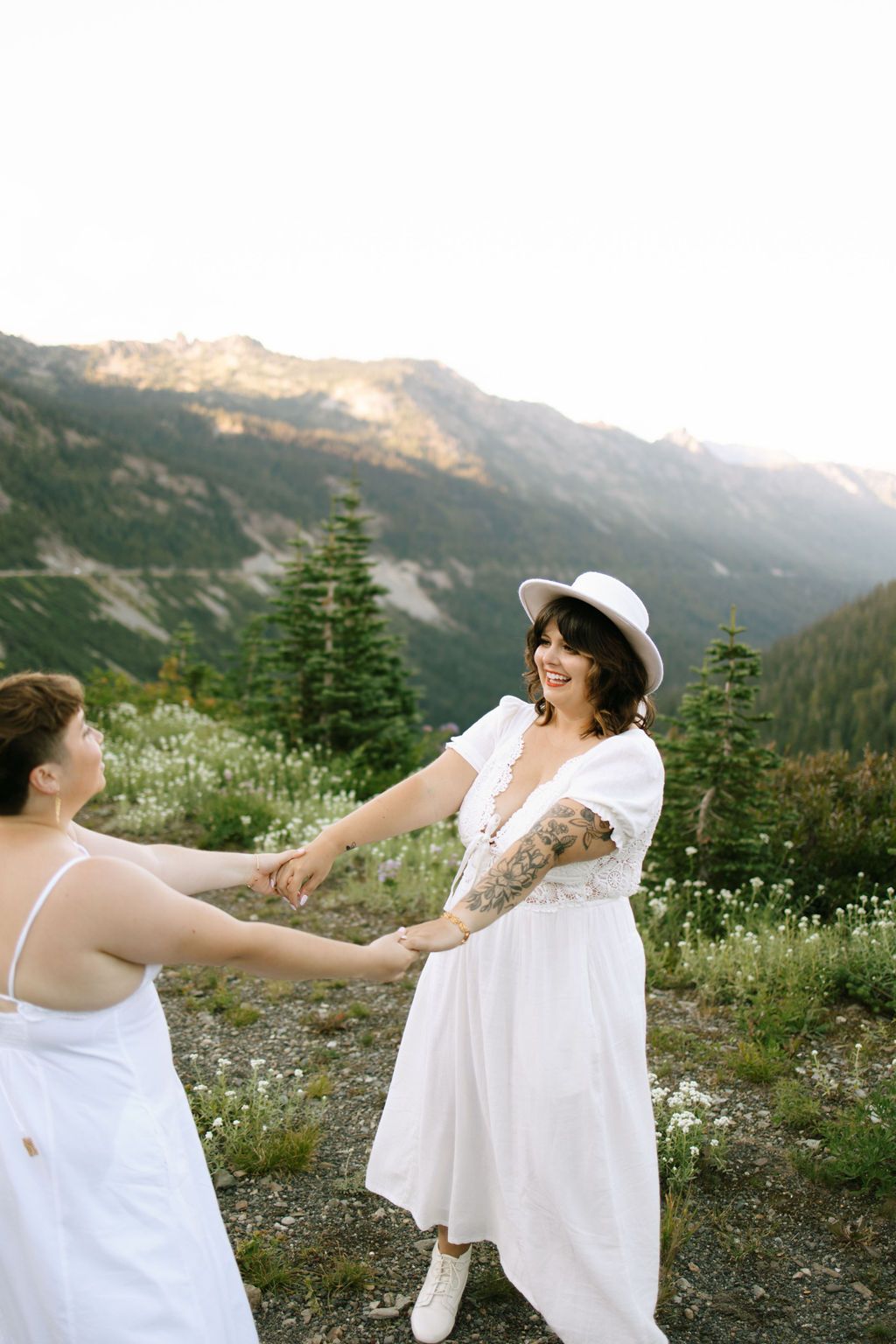 Two women in white dresses hold hands on a mountain meadow, smiling, with evergreen trees and peaks behind them.
