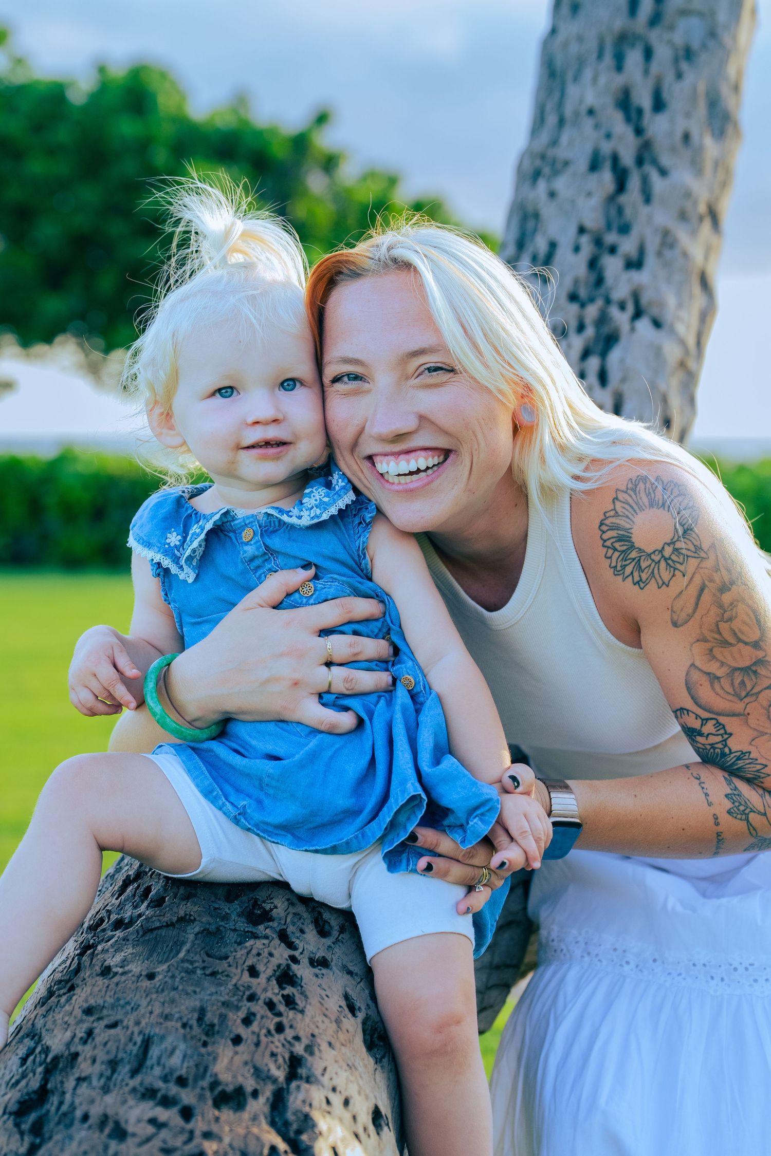 Smiling blonde woman with tattoos hugging a toddler girl in a blue dress outdoors near a palm tree.