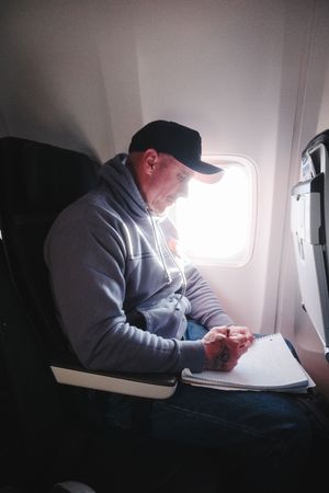 Man in gray hoodie and black cap writing in notebook while seated by airplane window during flight.