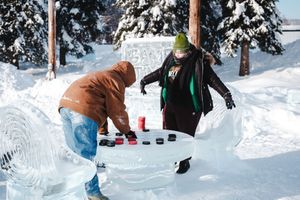 Two people playing a checker-like game on an ice table sculpture outdoors in a snowy winter setting.