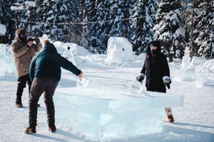 People examining large ice blocks during an outdoor winter ice sculpting event surrounded by snow-covered trees.