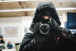 Photographer in black winter gear and face mask capturing photos with a professional camera indoors.