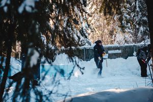 A person shoveling snow in a sunlit backyard surrounded by snow-covered trees in winter.
