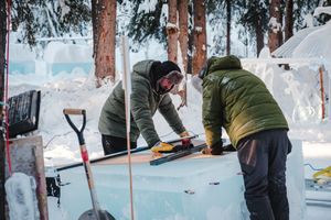 Two people in winter jackets working on carving a large ice block with tools in a snowy outdoor setting.