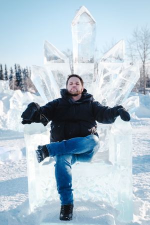 Man posing confidently on an ice throne sculpture outdoors in a snowy winter landscape.
