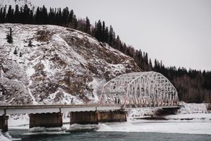 Steel arch bridge spans a snowy river valley with pine-covered mountains in winter landscape.