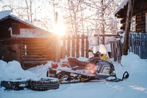 A snowmobile parked beside rustic wooden cabins bathed in golden sunset light during winter.