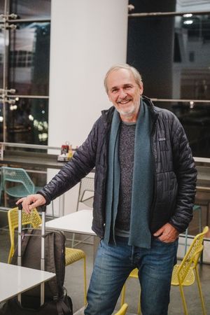 Smiling middle-aged man in a black Patagonia jacket and teal scarf standing in a modern cafe with yellow chairs.