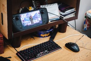 A Steam Deck handheld gaming console propped on a wooden desk beside a mechanical keyboard and mouse.