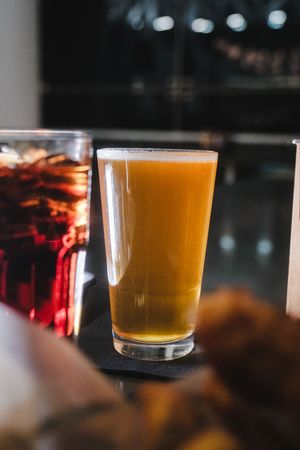 A pint glass of golden craft beer on a dark bar counter with a blurred red drink in the background.
