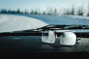 Frosted eyeglasses resting on a car dashboard with a snowy winter landscape visible through the windshield.