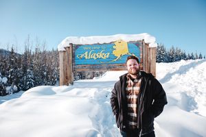 Man smiling in front of Welcome to Alaska sign surrounded by snow-covered trees on a sunny winter day.