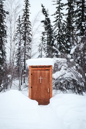 A rustic wooden outhouse with a cross on the door standing in a snowy forest in winter.