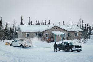 Two trucks parked outside a snow-covered house in a winter landscape with tall evergreen trees in the background.