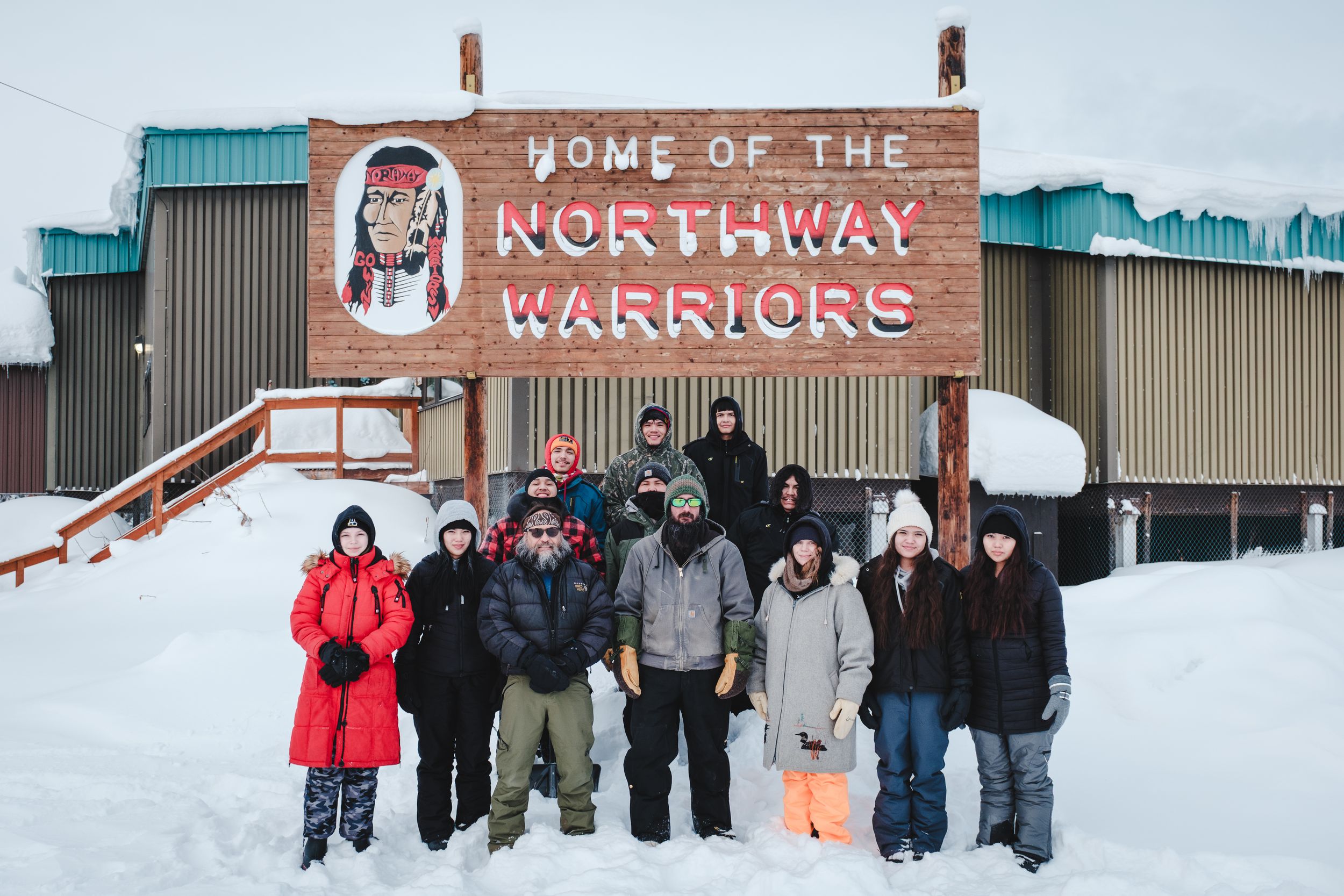 Group of people posing outdoors in winter gear in front of the Home of the Northway Warriors sign in snow.