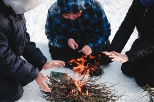 Three people warming their hands over a small campfire built on snow in winter.