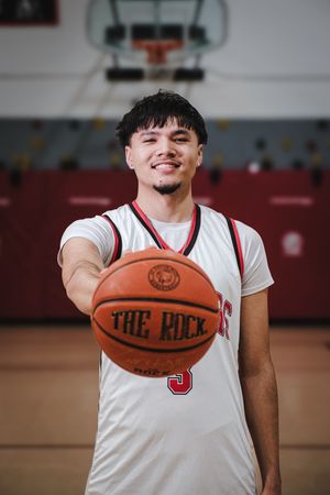 Smiling basketball player in white jersey holding The Rock basketball toward camera in indoor gym.