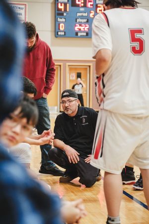 Basketball coach crouching on court giving team instructions during timeout with scoreboard showing 2:22 remaining.