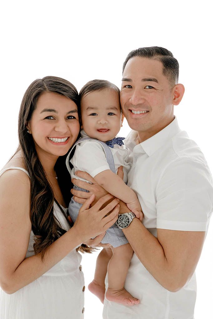 Smiling family posing together, parents in white holding their cheerful baby boy in a studio portrait near French Valley, California.